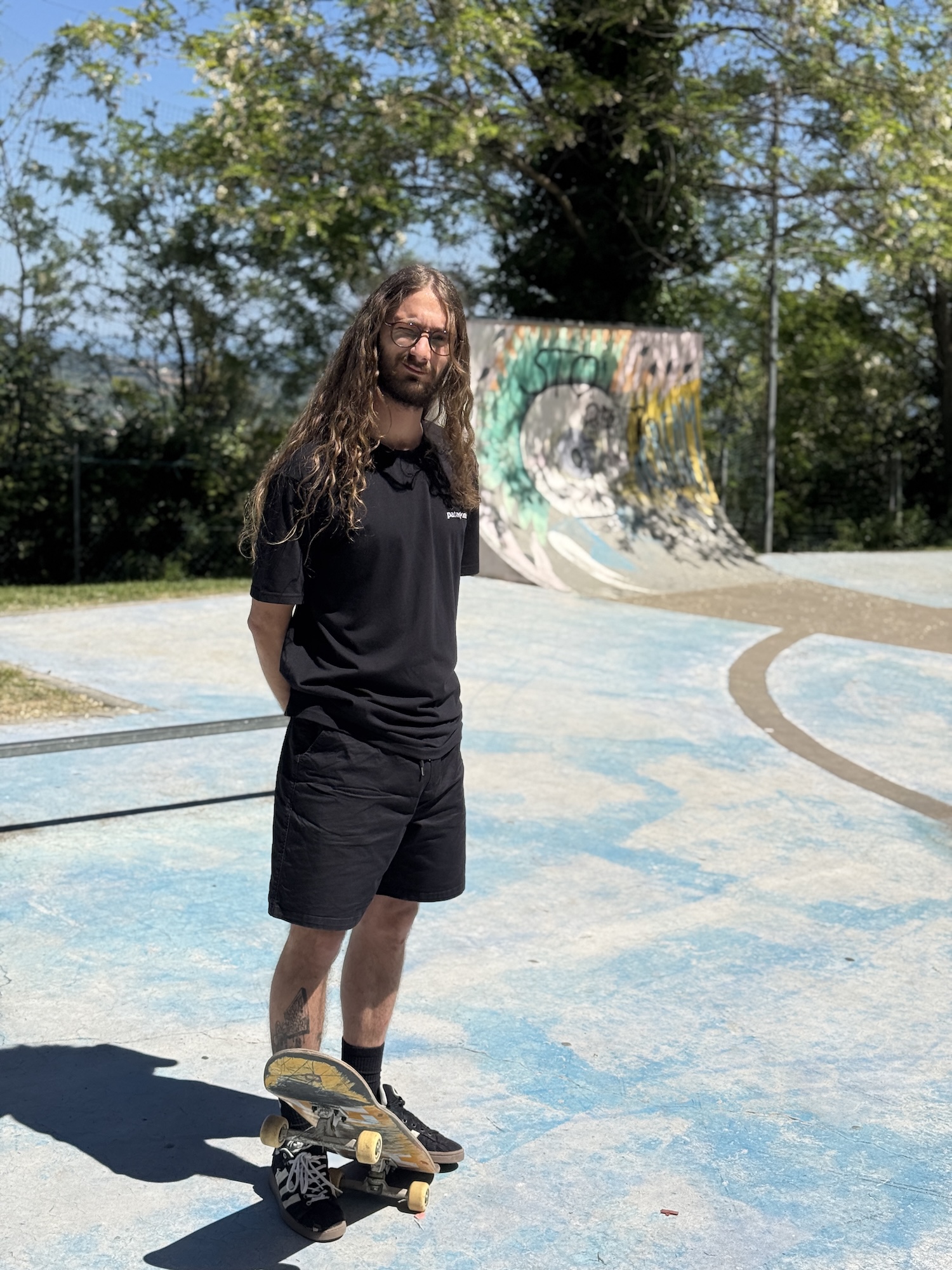 Bradipo, founder di Spot2Fakie, in piedi su uno skatepark con la sua tavola — capelli lunghi, t-shirt nera, look raw e autentico.